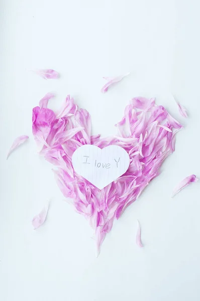 Heart made of pink peony petals on a white background. Around the heart, petals of a peony are casually scattered. In the center of the heart of the petals is a heart made of a leaf from a notebook with the inscription \