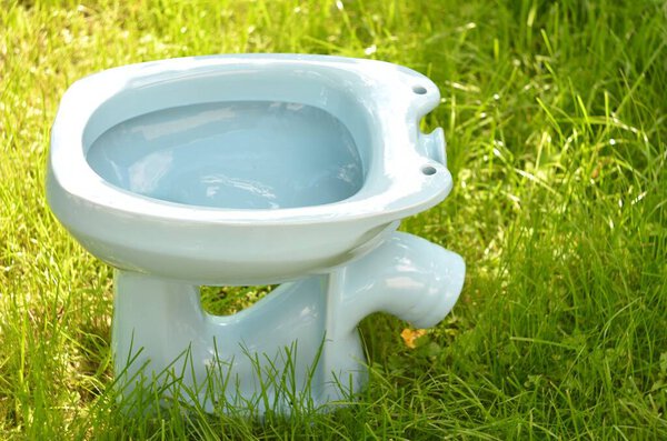 Туалет сделан в СССР в белом цвете. Вид сверху слева на фоне зеленой травы в солнечный день
.