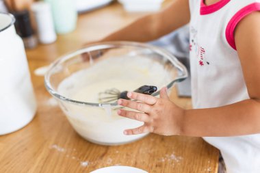 Küçük kız mutfakta krep için hamur hazırlıyor. Yemek hazırlama kavramı, arka planda beyaz mutfak. Gerçek hayatta sıradan bir fotoğraf serisi.