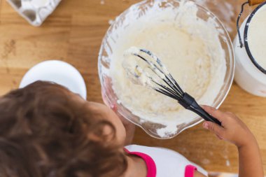 Küçük kız mutfakta krep için hamur hazırlıyor. Yemek hazırlama kavramı, arka planda beyaz mutfak. Gerçek hayatta sıradan bir fotoğraf serisi.
