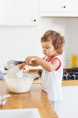 Küçük kız mutfakta krep için hamur hazırlıyor. Yemek hazırlama kavramı, arka planda beyaz mutfak. Gerçek hayatta sıradan bir fotoğraf serisi.
