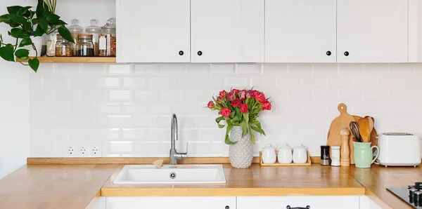 Tulips bouquet in vase standing on wooden countertop in the kitchen. Modern white u-shaped kitchen in scandinavian style.