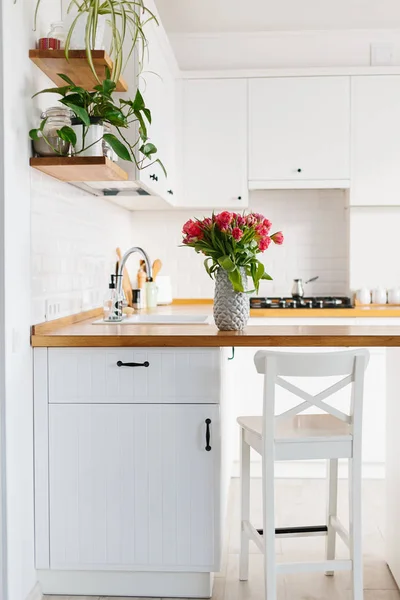 Tulips bouquet in vase standing on wooden countertop in the kitchen. Modern white u-shaped kitchen in scandinavian style.