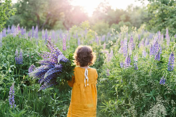 Чарівна кучерява маленька дівчинка в гірчичній лляній сукні з букетом квітів лаупіна — стокове фото