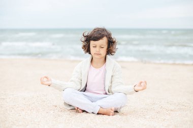 Küçük kız sahilde meditasyon yapıyor, yoga yapıyor, pastel renkler yapıyor.