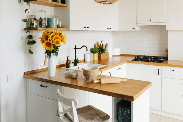 Tabaklar mutfak masasında, yemek yapmaya hazır. İskandinav tarzında beyaz, basit, modern mutfak, mutfak detayları, ahşap masa, masadaki ayçiçeği buketi.