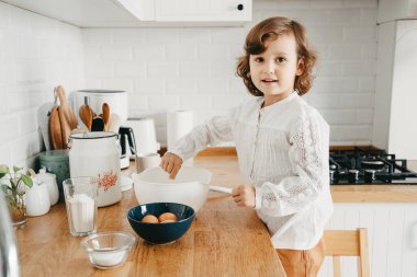 Küçük kız mutfakta krep için hamur hazırlıyor. Yemek hazırlama kavramı, arka planda beyaz mutfak. Gerçek hayatta sıradan bir fotoğraf serisi.