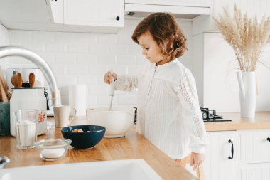 Küçük kız mutfakta krep için hamur hazırlıyor. Yemek hazırlama kavramı, arka planda beyaz mutfak. Gerçek hayatta sıradan bir fotoğraf serisi.