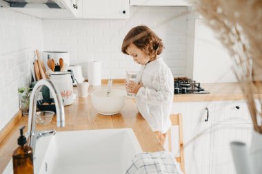 Küçük kız mutfakta krep için hamur hazırlıyor. Yemek hazırlama kavramı, arka planda beyaz mutfak. Gerçek hayatta sıradan bir fotoğraf serisi.