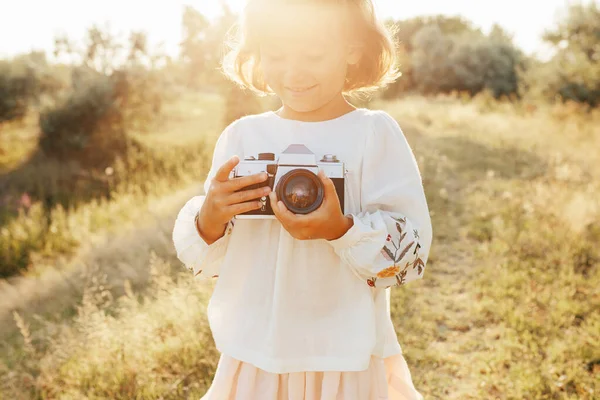 Little girl in embroidery blouse, rustic style dress, photographing with retro photo camera backlit summer sunset, positive vibes, rural simple life. Atmospheric image