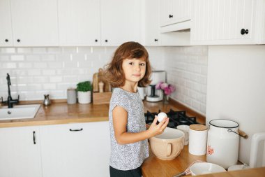 Çocuk kız pişirmek için hamur hazırlıyor - kasede yumurta kırıyor - ev yemeği. Yemek hazırlama kavramı, arka planda beyaz mutfak. Gerçek hayatta sıradan bir fotoğraf serisi.