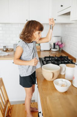 Çocuk kız mutfakta krep için hamur hazırlıyor. Yemek hazırlama kavramı, arka planda beyaz mutfak. Gerçek hayatta sıradan bir fotoğraf serisi.