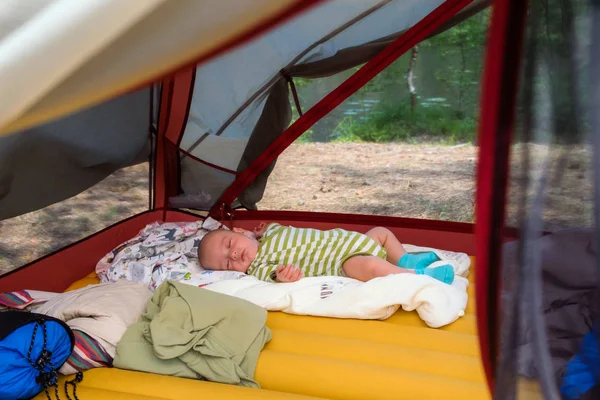 Küçük çocuk yaz gölüne çok uzak olmayan bir çadırın içinde yeşil çizgili giysiler içinde sırt ında uyuyor. — Stok fotoğraf