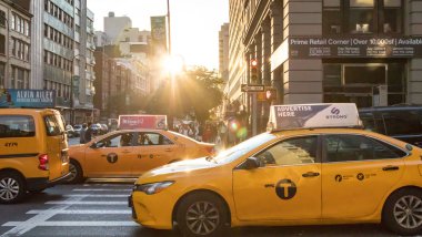 NEW YORK CITY, CIRCA 2018: Sarı taksi 5. Cadde 'de işlek bir kavşaktan geçiyor ve Manhattan siluetinin arka planında güneş ışığı parlıyor..