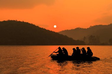 Group of soldiers help to row boat to target at twilight, mountain and sun in background. clipart