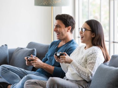 Yakışıklı beyaz adam ve güzel Asyalı kadın aşığı evde oturma odasında oturuyor ve birlikte oyun oynamak için joystick kullanıyorlar mutluluk ve neşeyle. Çeşitli çiftler ve ev konsepti..