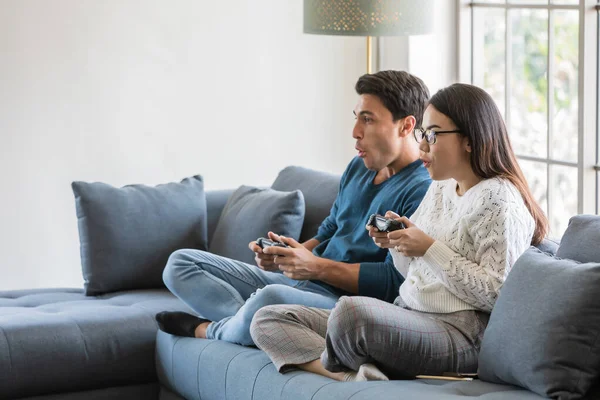 Yakışıklı beyaz adam ve güzel Asyalı kadın aşığı evde oturma odasında oturuyor ve birlikte oyun oynamak için joystick kullanıyorlar mutluluk ve neşeyle. Çeşitli çiftler ve ev konsepti..