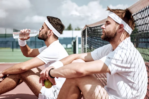 Relaxing after good game. Cheerful men at the tennis net and drinking water while both sitting on tennis court