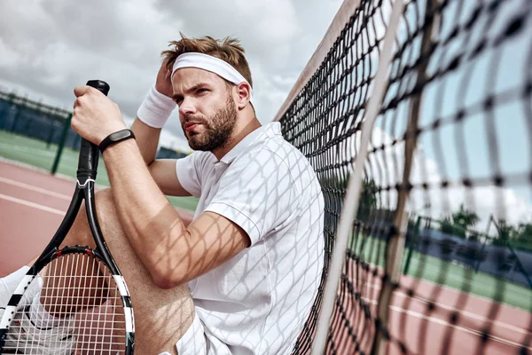 Relaxing after good game. Cheerful man at the tennis net resting sitting on tennis court