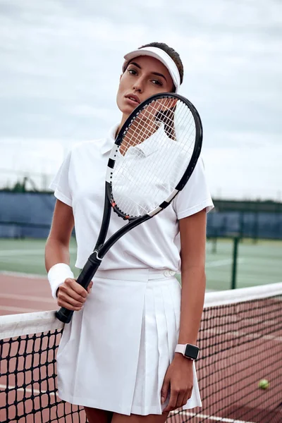 Portrait of fashionable woman in white clothing and cap with tennis racket posing at tennis net on court