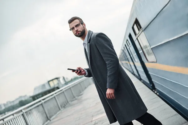 Business trip. Young businessman standing near railway holding smartphone looking aside pensive