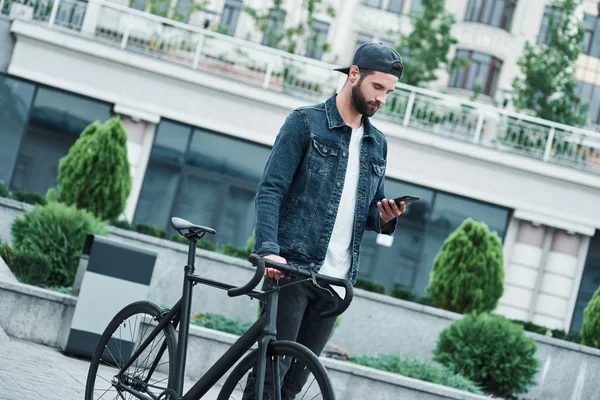 Outdoors leisure. Young stylish man walking on city street with bicycle browsing internet on smartphone concentrated