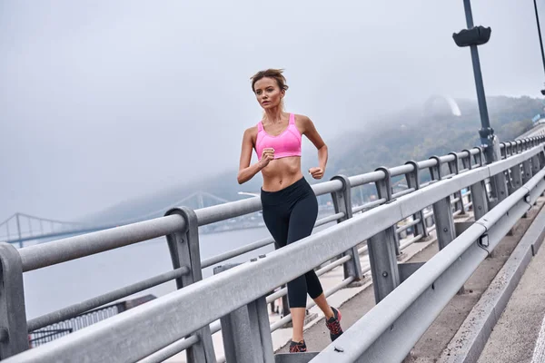 Always in great shape. Full length of young womanin sports clothing exercising while jogging on the bridge outdoors