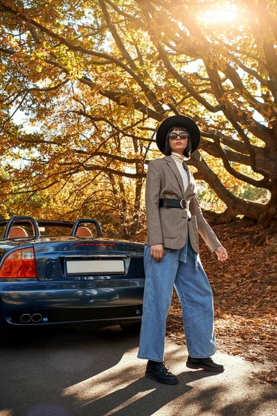 Attractive young woman posing near cabriolet. She wears black hat and sunglasses. Stylish autumn.