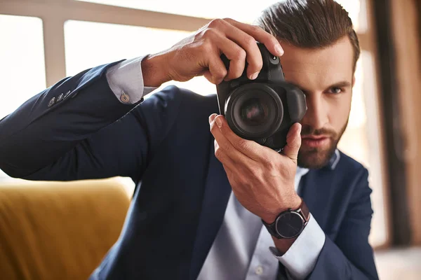 Young stylish businessman with camera in his hands taking pictures while sitting on sofa at modern office