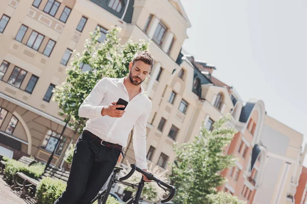 Make the most of yourself. Young brown-haired man standing in the park with a bicycle and looking at his phone