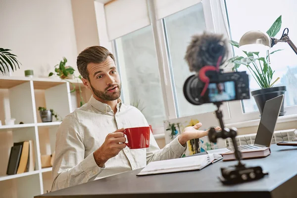 Creating content. Cheerful male blogger making a new video for his vlog and drinking a tea while sitting indoors.