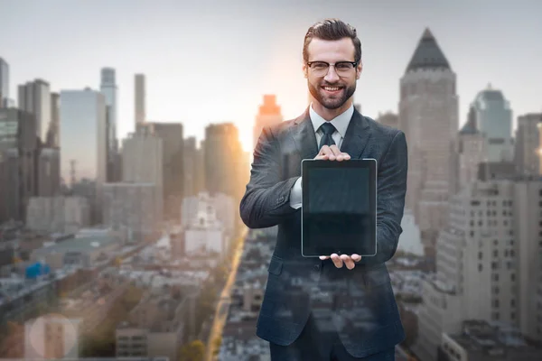 Making business easier. Portrait of cheerful young man in suit showing digital tablet and smiling while standing against of morning cityscape background