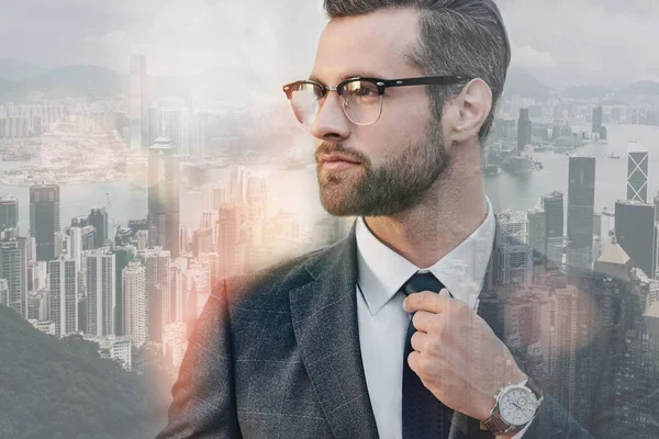 Confident business expert. Young bearded businessman in glasses adjusting necktie while standing outdoors with cityscape on the background