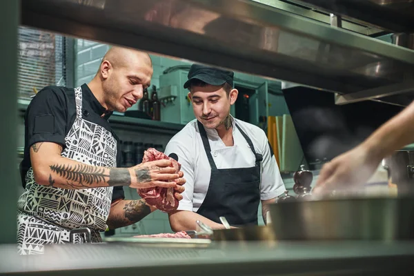 How to choose the right meat Professional chef in apron and with tattoos on his arms showing a red meat to his assistant