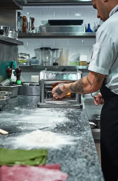 Making pasta. Vertical image of chefs hands with tattoos rolling a dough through pasta machine in restaurant kitchen