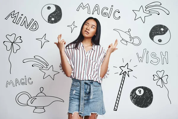 I know magic trick. Cute asian young woman in casual wear crossing fingers and making a wish while standing against grey background with hand drawn magic tools on it