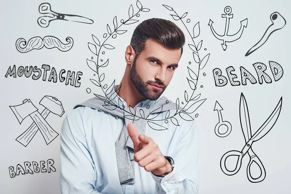 Right choice. Charming bearded man with perfect hairstyle gesturing and looking at camera while standing against grey background with doodles on the theme of barber shop.