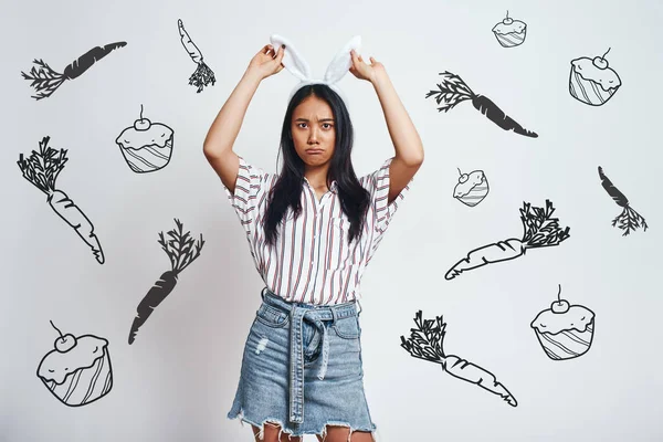 Bored bunny. Cute young Asian woman in bunny ears looking at camera and making a face while standing against grey background with hand drawn carrots and cupcakes on it