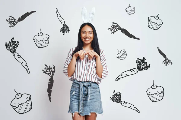 Funny bunny. Cute young Asian woman in bunny ears and casual wear is smiling while standing against grey background with hand drawn carrots and cupcakes on it