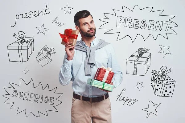 Buying gifts in advance. Good looking and smiling man is holding colorful gifts for friends while standing against grey background with different doodle illustrations on it