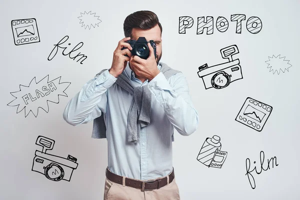 Do what you love. Young bearded man in blue shirt taking a picture on digital camera while standing against grey background with different doodle illustrations on it. Hobby