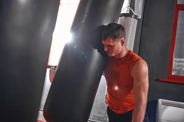 Tired after workout. Tired young athlete in sports clothing looking away after hard training on heavy punch bag in boxing gym