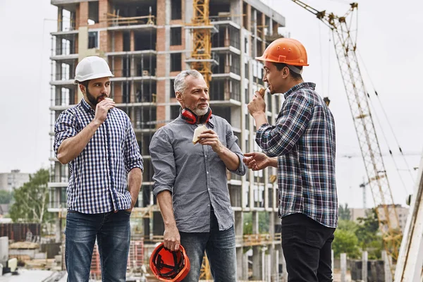 Lunch time. Group of builders in working uniform are eating sandwiches and talking while standing at construction site.