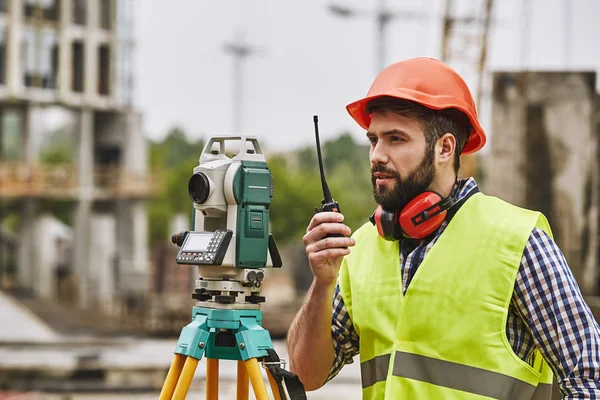 Precise measurements. Surveyor engineer in protective wear and red helmet using geodetic equipment and talking by walkie talkie while standing at construction site. Professional equipment.