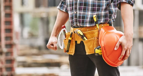 Safe work. Cropped photo of male professional builder with construction tools holding a safety red helmet while standing outdoor of construction site