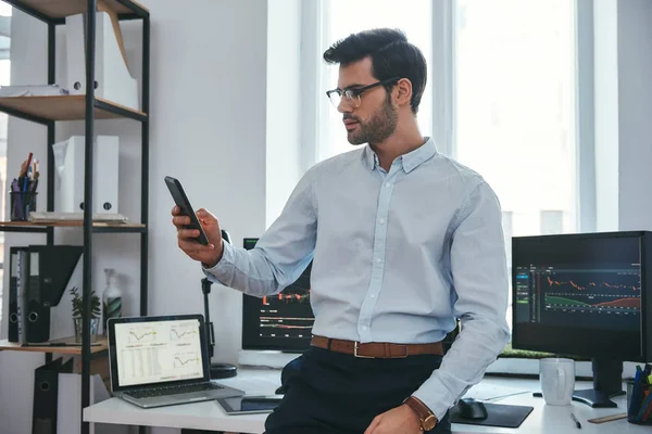Stock exchange. Concentrated young trader or businessman in formal wear and eyeglasses is looking at trading reports on his smartphone for investment purpose while working at his office.