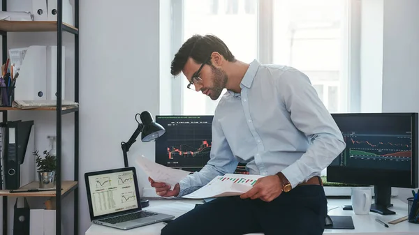 Busy working day. Young successful trader or businessman in formal wear and eyeglasses sitting at modern office in front of computer screens with charts and reading market report