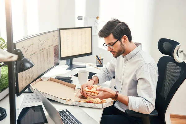 I love pizza Young bearded businessman or trader in eyewear and formal clothes is eating hot pizza while sitting in his modern office.