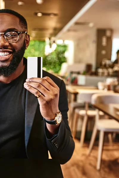 Price is what you pay. Value is what you get. Smiling african american businessman holds his credit card, looking at the camera while standing in the cafe
