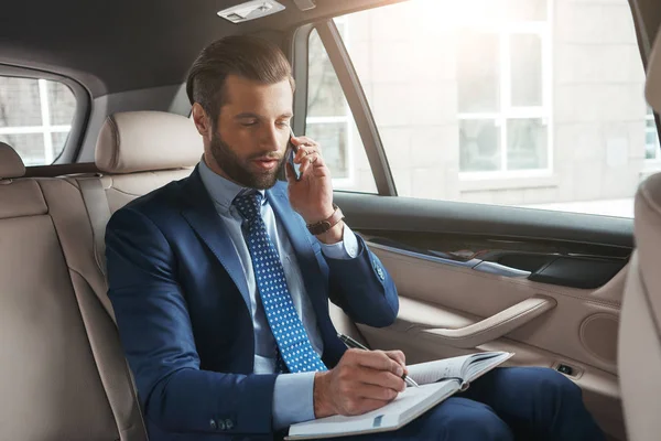 Important talk with client. Busy young businessman in formal wear writing something in his notebook and talking with client by smartphone while sitting on the back seat of his car